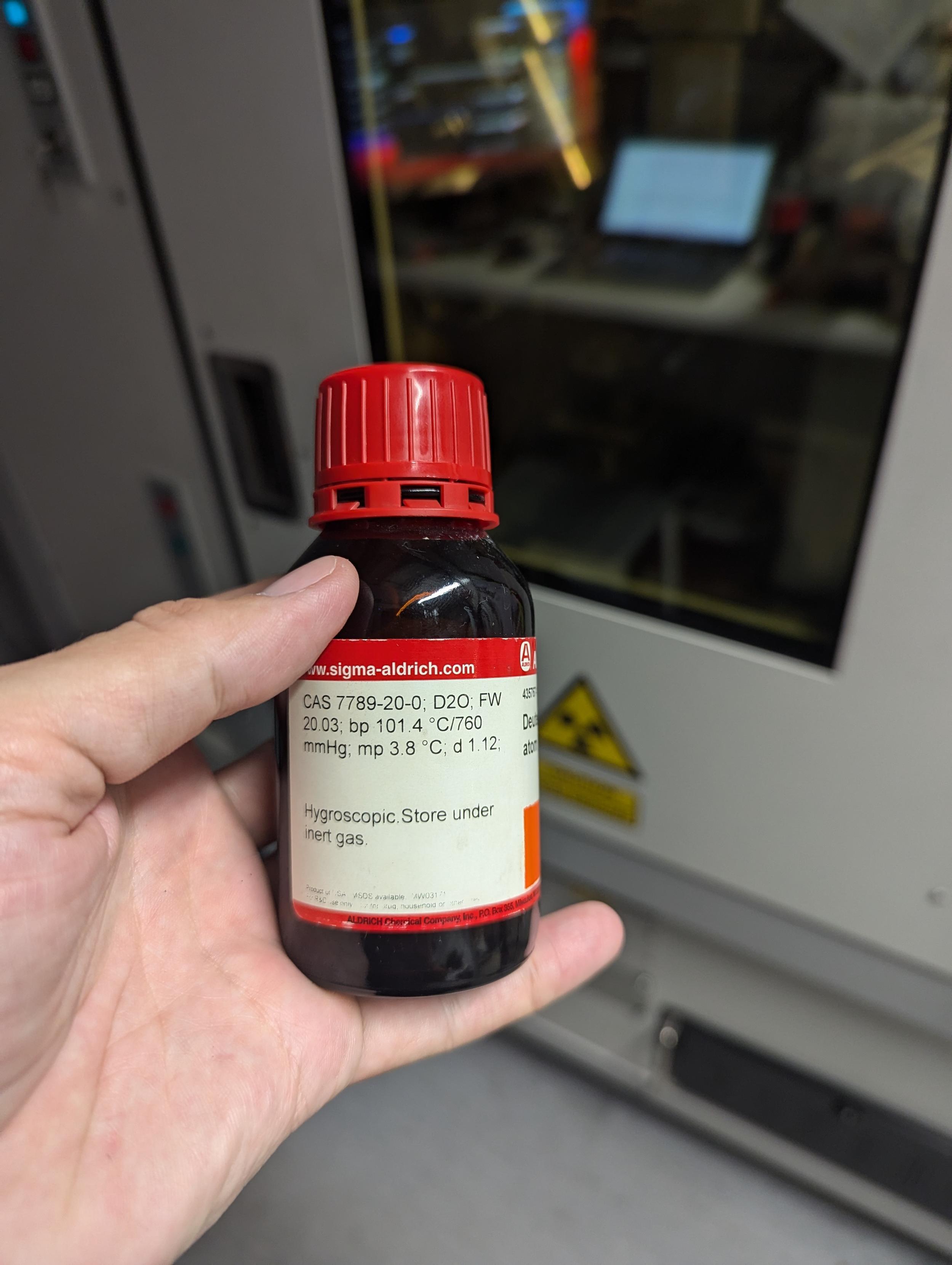 A hand holding a brown bottle with a red cap and a white label.  The label reads 20.03; BP 101.4 MP 3.8 C. D 1.12  Hydroscopic store under inert gas